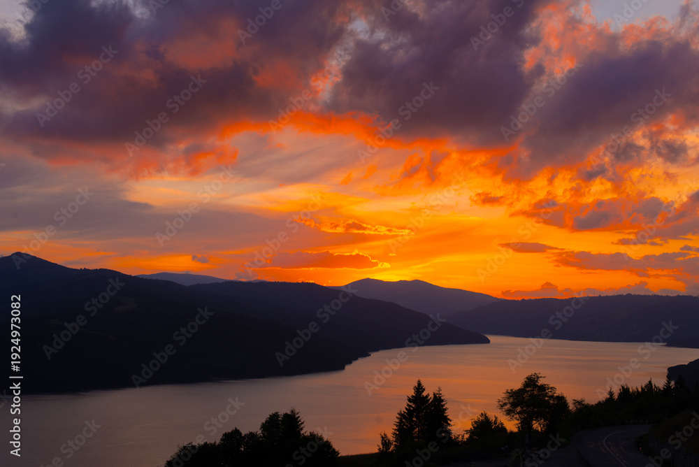 Naklejka premium A vibrant sunset casts warm colors over a serene lake nestled between towering mountains, creating a peaceful atmosphere in nature's embrace.