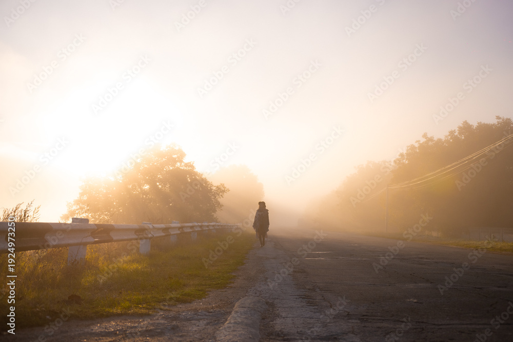 Fototapeta premium A woman walks through thick fog on a deserted road as the sun rises in the distance, casting soft light on the serene scene of nature awakening.