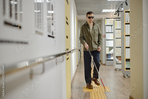 Caucasian young adult man wearing sunglasses walking through library corridor using cane for navigation, visually impaired individual confidently moving along tactile paving