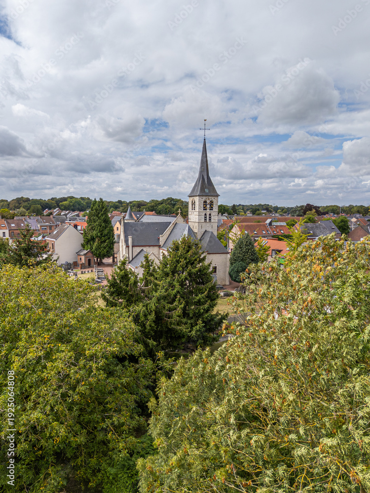 Fototapeta premium Aerial, bird view church of Eppegem village, a small rural village in the countryside of Belgium. deciduous trees in foreground. Houses around the church. Cloudy autumn day. Beautiful rural town. Natu