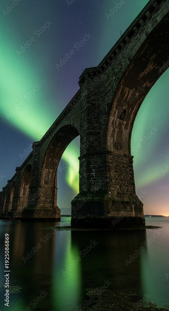 Fototapeta premium Low angle of stone bridge with arches in water reflecting aurora borealis lights against starry sky, representing travel, engineering and nature