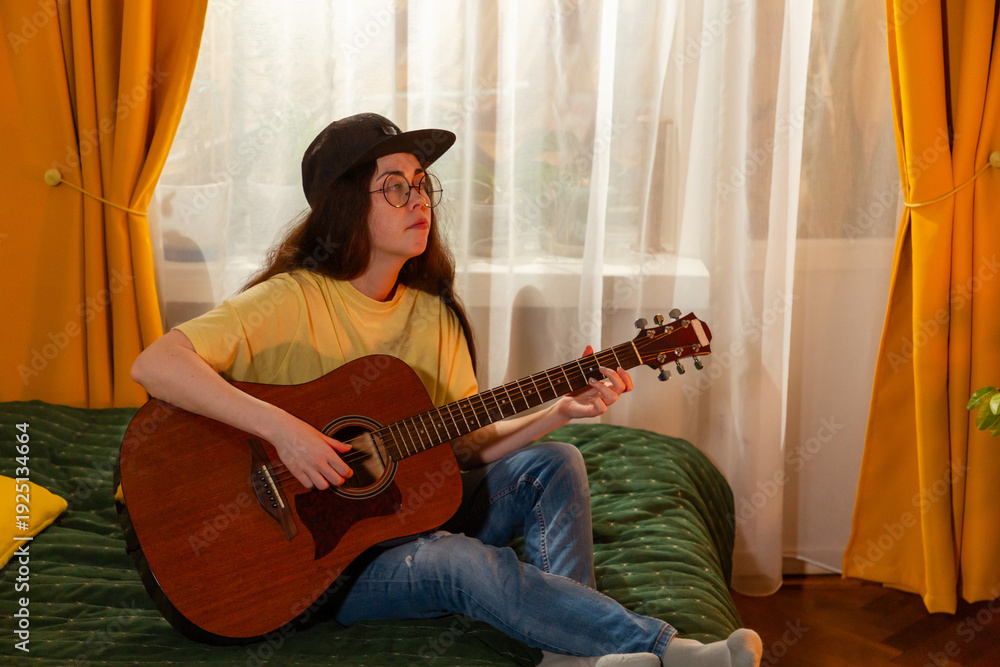 © _KUBE_ - Caucasian woman student plays guitar in bedroom. She sits on bed during music practice. Creative education theme. Mid shot