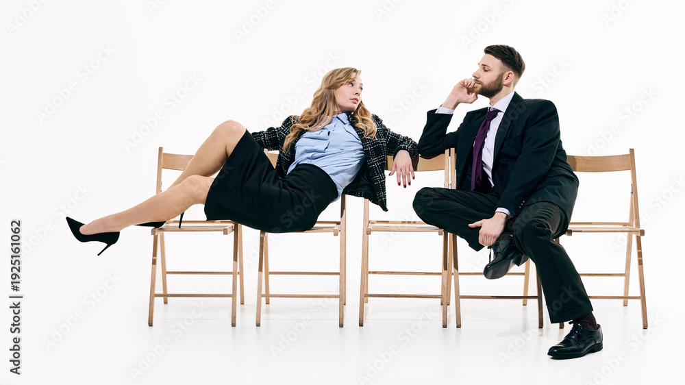 Obraz premium Elegant business couple posing across wooden chairs in white space. Concept of power dynamics, office relationships and contemporary corporate fashion campaign with subtle tension.