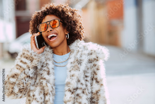 Latin american woman with afro hairstyle, laughing while talking on mobile phone wearing a fur coat in the city.