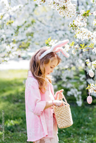 Outdoor Easter celebration, childhood joy and seasonal tradition. Little girl with bunny ears holding decorative Easter egg and wicker basket under blooming spring tree.