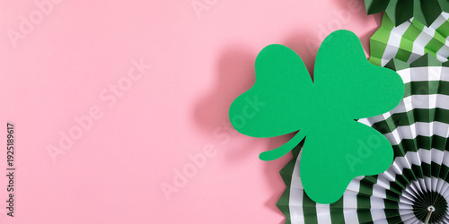 St. Patrick's Day celebration banner with green paper shamrock and decorative fans on pastel pink background. Minimalist Irish holiday flat lay with copy space for greeting text or advertising.