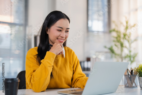 Photos Smiling young Asian millennial woman working remotely, typing on a laptop comput