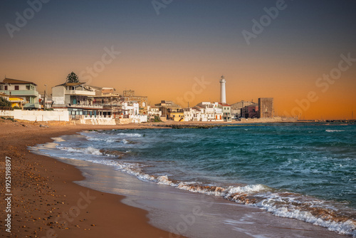 Waves breaking along the sandy beach in Punta Secca, Sicily, under a warm sunset sky
