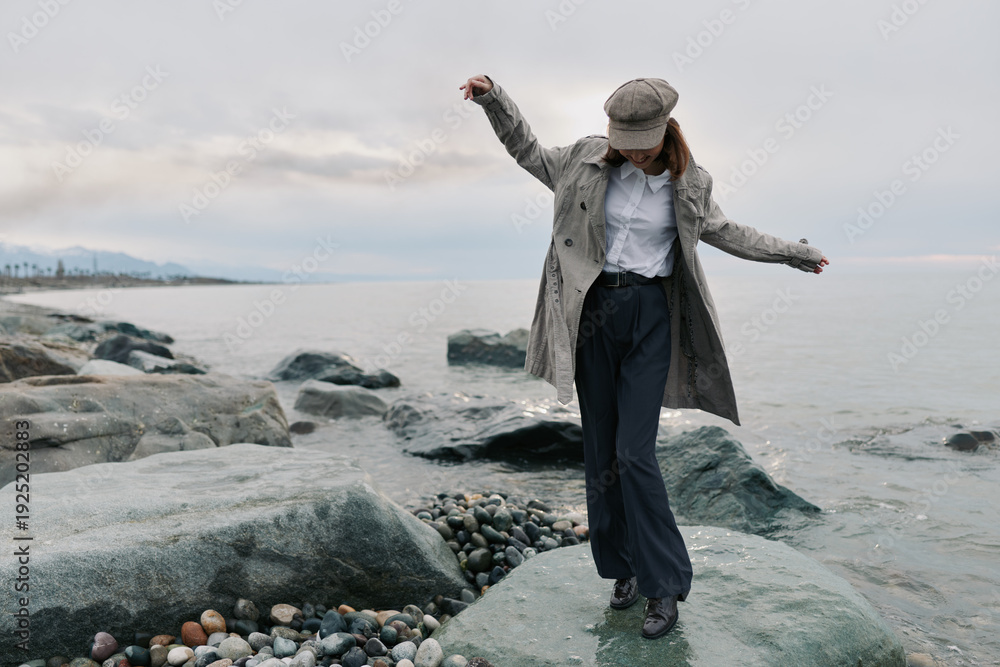 Obraz premium Woman in stylish coat and cap balances on rocky shore near calm sea under cloudy sky. Outdoor fashion, leisure, and nature concept with casual autumn outfit and relaxed mood.