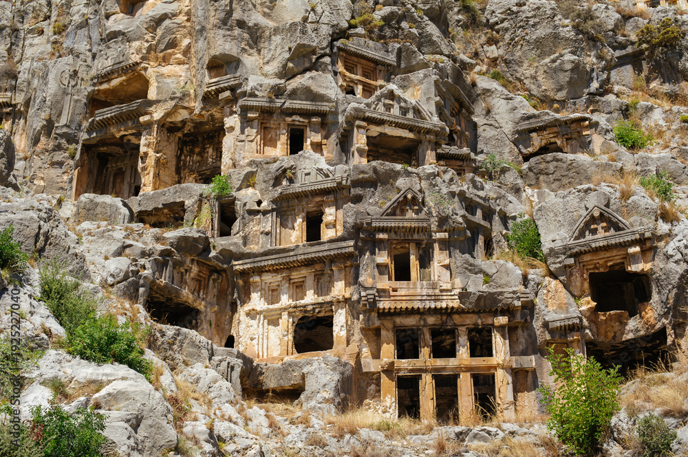 Naklejka premium A horizontal shot showcasing the detailed architectural facades of ancient Lycian tombs carved into the vertical limestone cliffs at Myra in Demre, Turkey.
