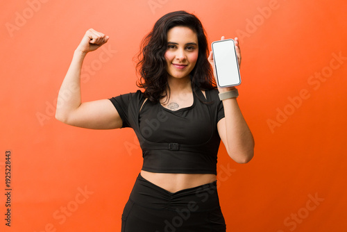 Obraz na plátně Empowered woman showing bicep and smartphone with blank screen, promoting fitnes