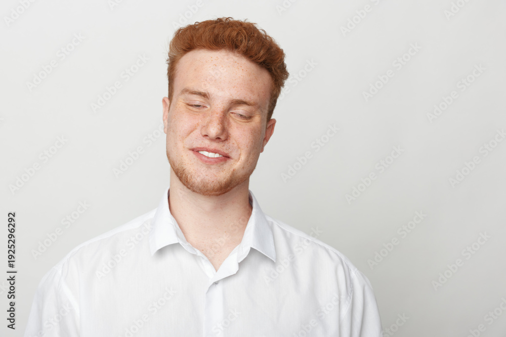 Obraz premium Young man with red hair and beard smiles gently with closed eyes wearing white shirt on light background. Concept of happiness, relaxation, and natural expression.