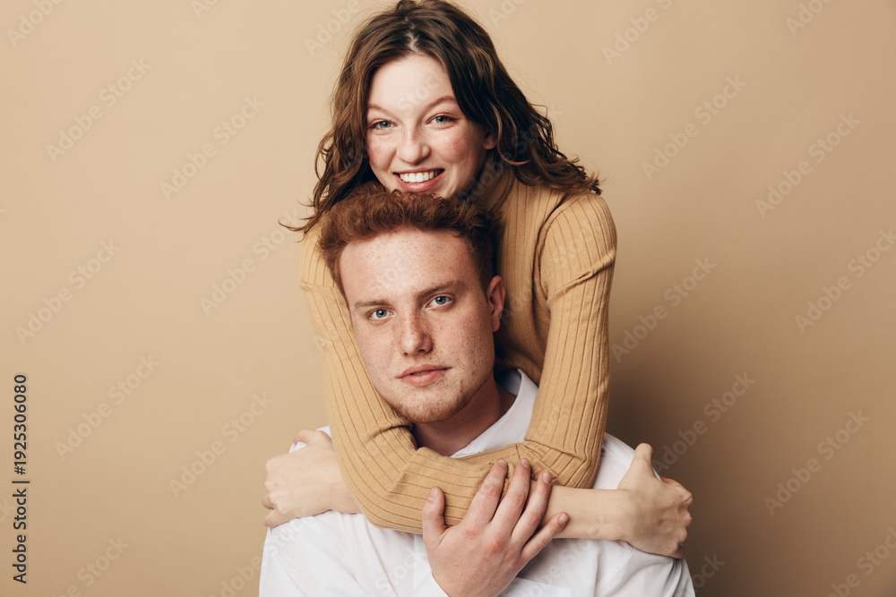 Fototapeta premium Smiling woman with curly hair hugs serious man from behind on beige background. Young couple shows affection and connection in casual clothing. Concept of love, relationship, and friendship.
