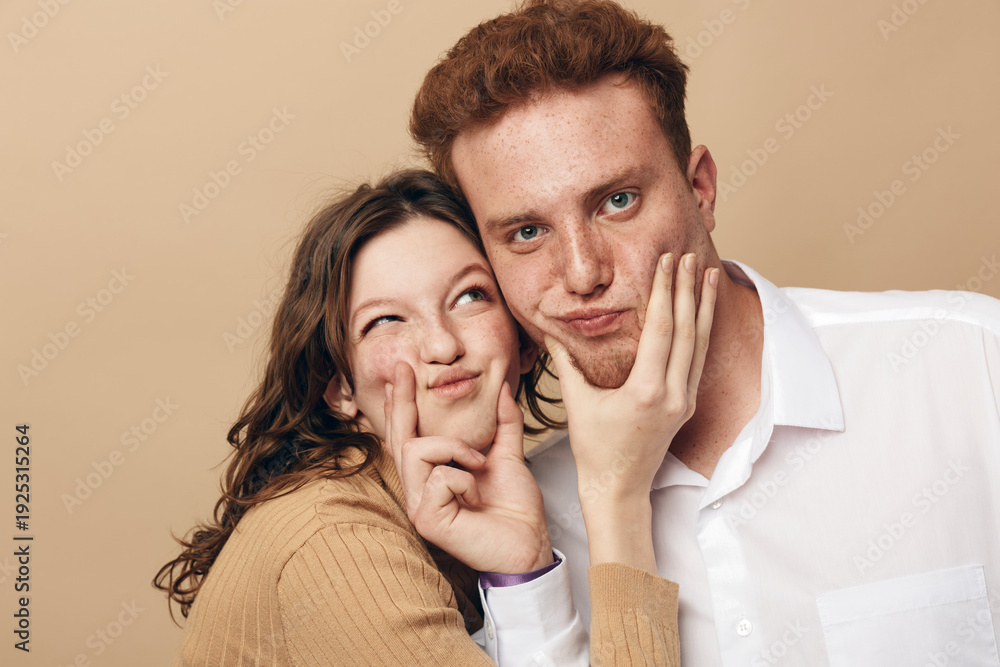 Fototapeta premium Young woman playfully pinches young man’s cheeks while both smile and pose against beige background. Concept of friendship, fun, affection, and casual lifestyle in studio portrait.