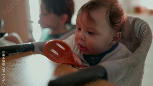 Quadro em tela Baby holding orange spoon with wide-eyed expression, joyful sibling in backgroun