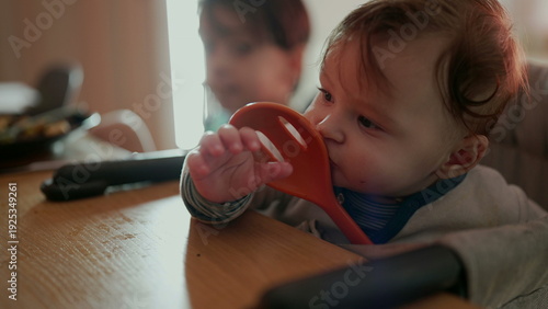 Bilde på lerret Baby holding orange spoon with wide-eyed expression, joyful sibling in backgroun