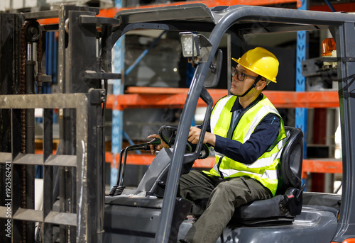 An adult Asian male warehouse worker, wearing a helmet and safety vest, operates a forklift in an industrial warehouse.