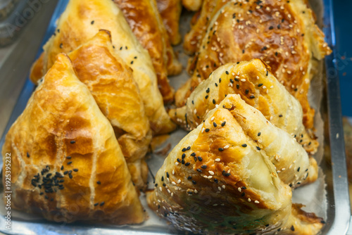Homemade golden uzbek samsa pastries. Freshly baked triangular samosas with black and white sesame seeds on top. Delicious oriental puff pastry pies arranged on a tray in a bakery.
