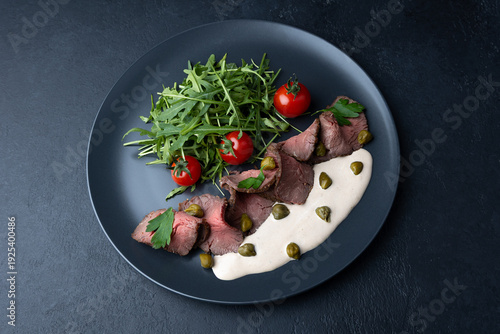 Vitello Tonnato with arugula, cherry tomatoes, capers, and parsley. A popular Italian appetizer made with veal and a tuna-anchovy sauce. Selective focus, black background, close-up.