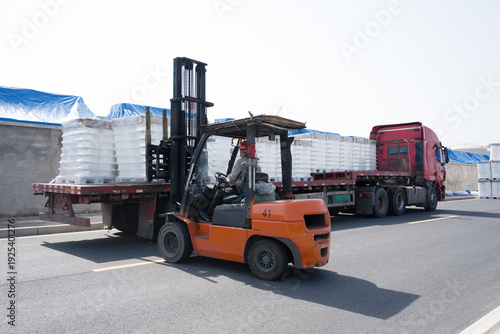 Tableau sur toile Orange forklift loading white bagged goods onto flatbed truck at industrial site