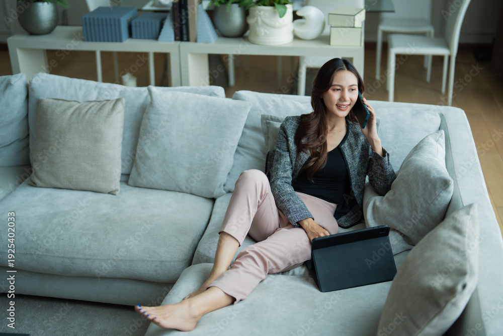 Obraz premium Woman in smart casual attire comfortably working from home, making a business phone call while sitting on a sofa with a tablet beside her, representing flexible modern work
