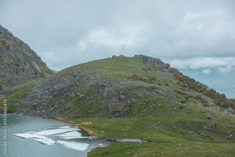 Fototapeta premium Gloomy view to turquoise lake near green hill in hanging valley against misty hills in poor visibility in low clouds. Ice floes float in upland lake in mountain cirque against foggy rainy vastness.