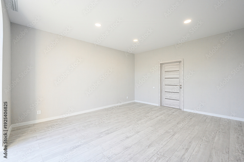 Obraz premium A spacious, empty room with light grey walls, wooden-style flooring, a light wood door, and recessed ceiling lights, ready for furnishing