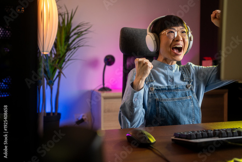 Fotografie Pretty and Excited Asian Gamer Girl in Headphones is Playing First-Person Shooter Online Video Game on Her Computer