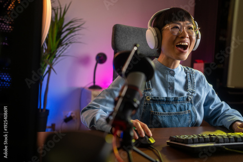 Fotografie Pretty and Excited Asian Gamer Girl in Headphones is Playing First-Person Shooter Online Video Game on Her Computer