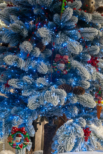 Frosted Christmas tree with blue lights and festive ornaments. Closeup of decorated snowy Christmas tree with glowing blue lights red berries pinecones and whimsical ornaments.