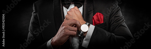 Foto A man in a formal black tuxedo and bow tie adjusts a stylish wristwatch with a striped strap against a dark background