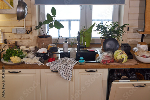 Messy modern kitchen after cooking with dirty dishes, fresh vegetables and cluttered countertop. Open dishwasher and black sink in realistic home interior. Everyday domestic life and household chaos 