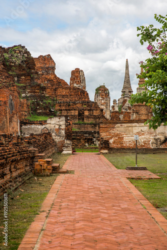 Ancient ruins in Ayutthaya, Thailand. A red-brick pathway leads the viewer's eye toward a collection of diverse historical structures, including bell-shaped chedis and a tall, weathered prang.