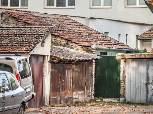 Cluster of weathered garages and rustic outbuildings with sloping tiled roofs and wooden doors, tucked inside an urban residential courtyard with parked cars and signs of age and renovation needs.