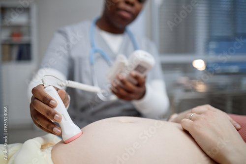 Black female doctor performing ultrasound scan on pregnant Caucasian woman, focusing on abdomen while holding medical device, healthcare professional conducting prenatal examination