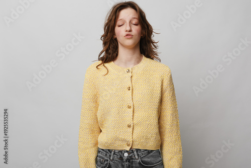 Fotografie Young woman in yellow knitted cardigan stands with closed eyes and relaxed expression against grey background
