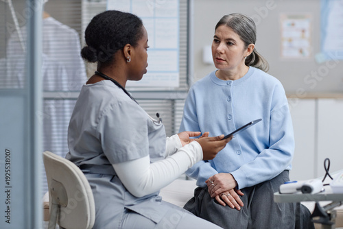 Black female nurse discussing medical information with middle aged Caucasian woman in clinical setting, nurse holding digital tablet while patient listening attentively during consultation