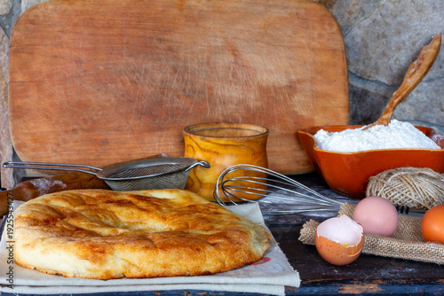 Rustic still life with fresh baked flatbread, flour, eggs, and baking utensils on a wooden table.