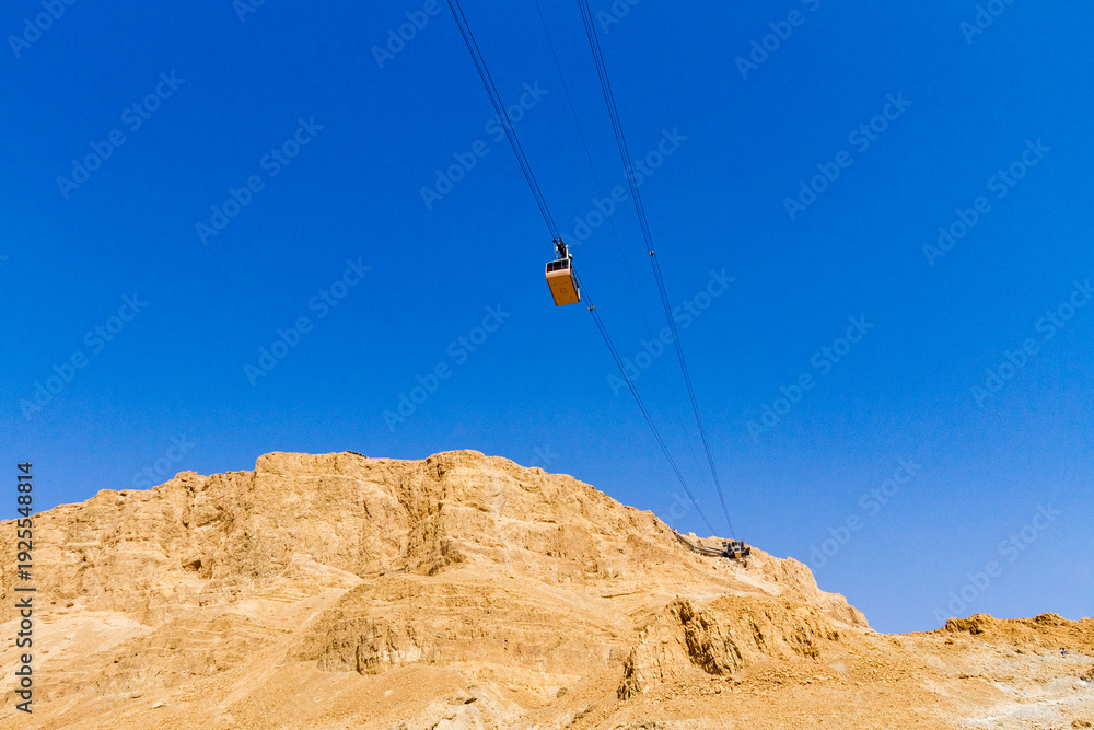 Obraz premium Cable car ascending to Masada fortress in Judean Desert, Israel. Ancient historic site near Dead Sea under bright blue sky.