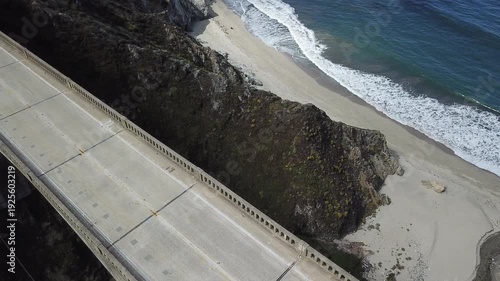 Cinematic aerial drone view over Bixby Creek Bridge. A soaring perspective of the iconic arch as it spans a deep canyon, where rugged Big Sur cliffs meet the crashing white surf of the Pacific Ocean.