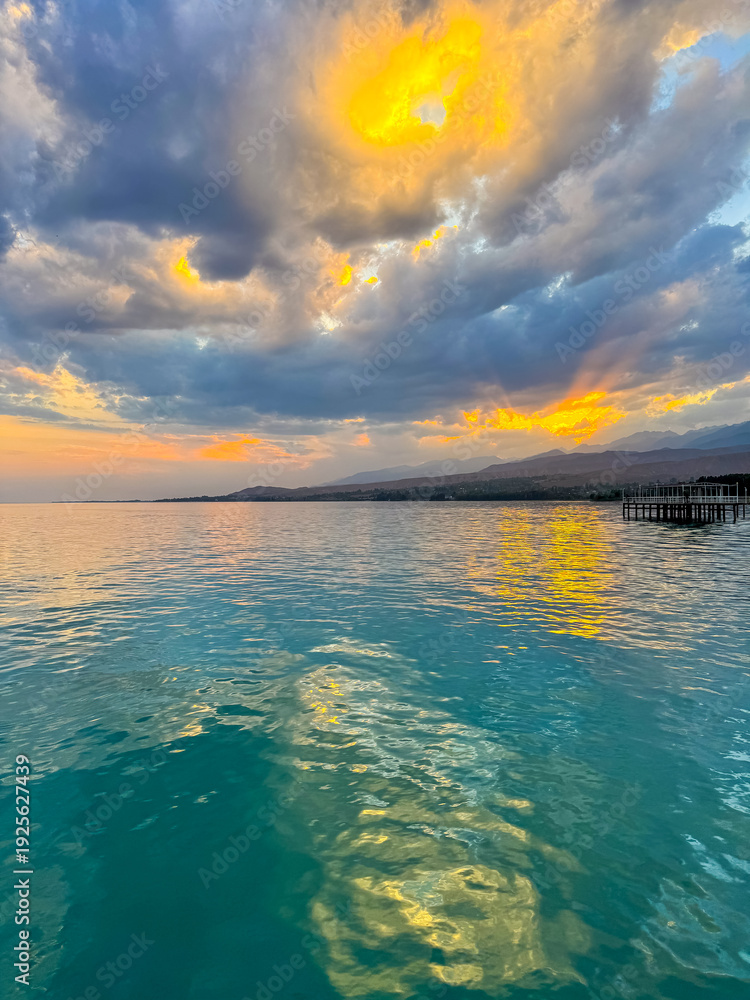 Fototapeta premium Sunset at Issyk Kul Lake in Kyrgyzstan. Bright sun breaking through clouds reflecting in clear turquoise water.