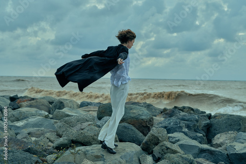 Foto Woman in a flowing cape stands on jagged rocks by the sea, wind lifting the fabr
