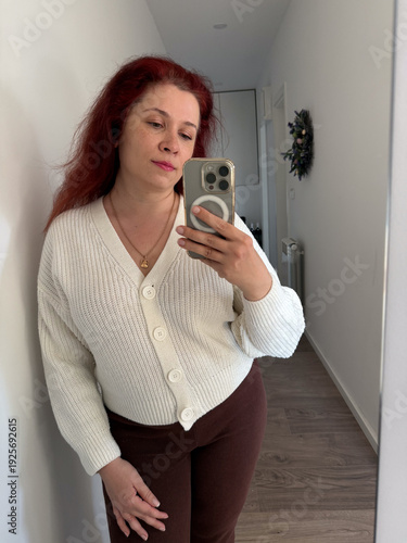 Woman taking mirror selfie in bright hallway. Casual outfit, personal style, everyday fashion, confidence concept