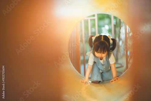 Happy little Asian girl playing in tunnel at outdoor playground, cheerful child crawling through play equipment in public park, concept of childhood development, active lifestyle, and learning