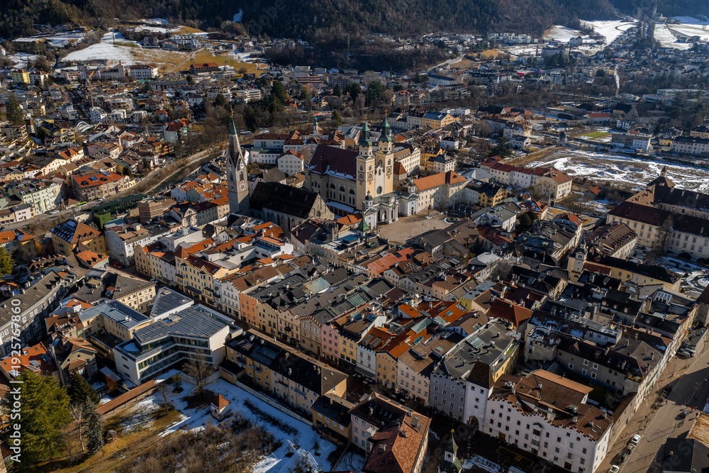 Fototapeta premium Aerial view of Brixen, Italy, shows Brixen Cathedral twin towers, Gothic bell tower, pastel facades, red roofs, cathedral square, winter hills, and long shadows.