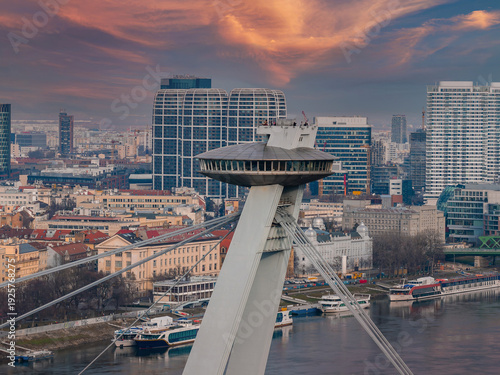 Foto Aerial scene in Bratislava, Slovakia, shows SNP Bridge UFO deck, asymmetrical pylon and cables, Danube River with docked boats, towers and historic riverside buildings at sunset