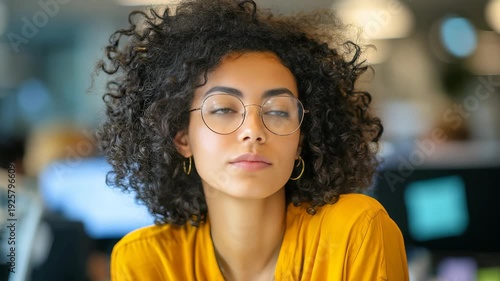 Portrait stylish woman with natural curls, round glasses, dressed in vibrant yellow shirt, sitting in contemporary workspace. Atmosphere suggests innovation, ambition, professionalism