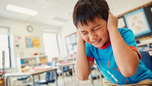 Distressed Asian boy covering ears in school classroom. Student experiencing sensory overload or loud noise. Child feeling anxious and overwhelmed. Mental health
