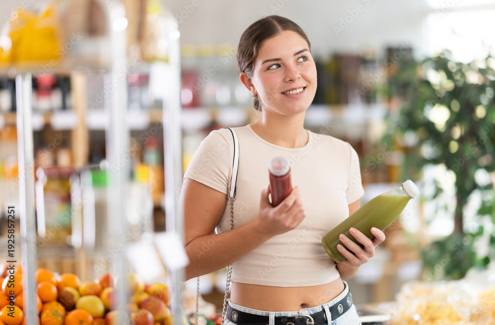 Naklejka premium Young woman buyer chooses red and green smoothie in bottle in grocery store