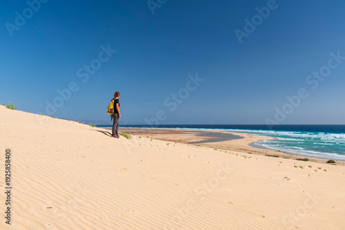 Photos Woman hiking on sand dunes overlooking a vast beach in Fuerteventura, Canary Islands, Spain, enjoying panoramic ocean views under a clear blue sky